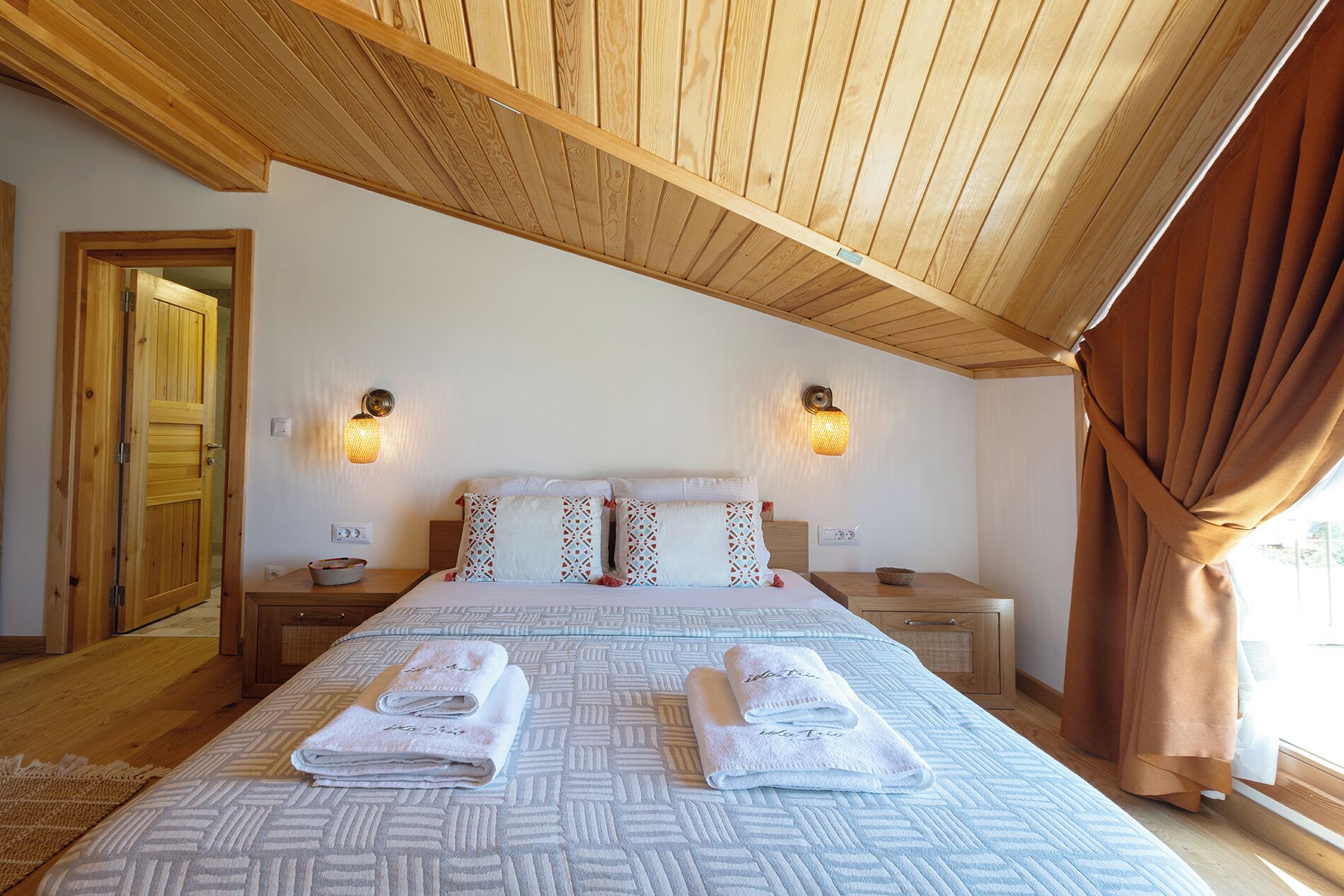 A hotel room with a wooden ceiling, a bed with patterned bedding, two bedside tables, and a window with curtains.