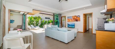 A modern living room with a sofa, a dining table with chairs, a ceiling fan, and a kitchen area with a refrigerator.