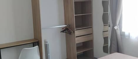 Une chambre à coucher comprenant un lit, un bureau, une chaise, une armoire et un ventilateur sur pied.