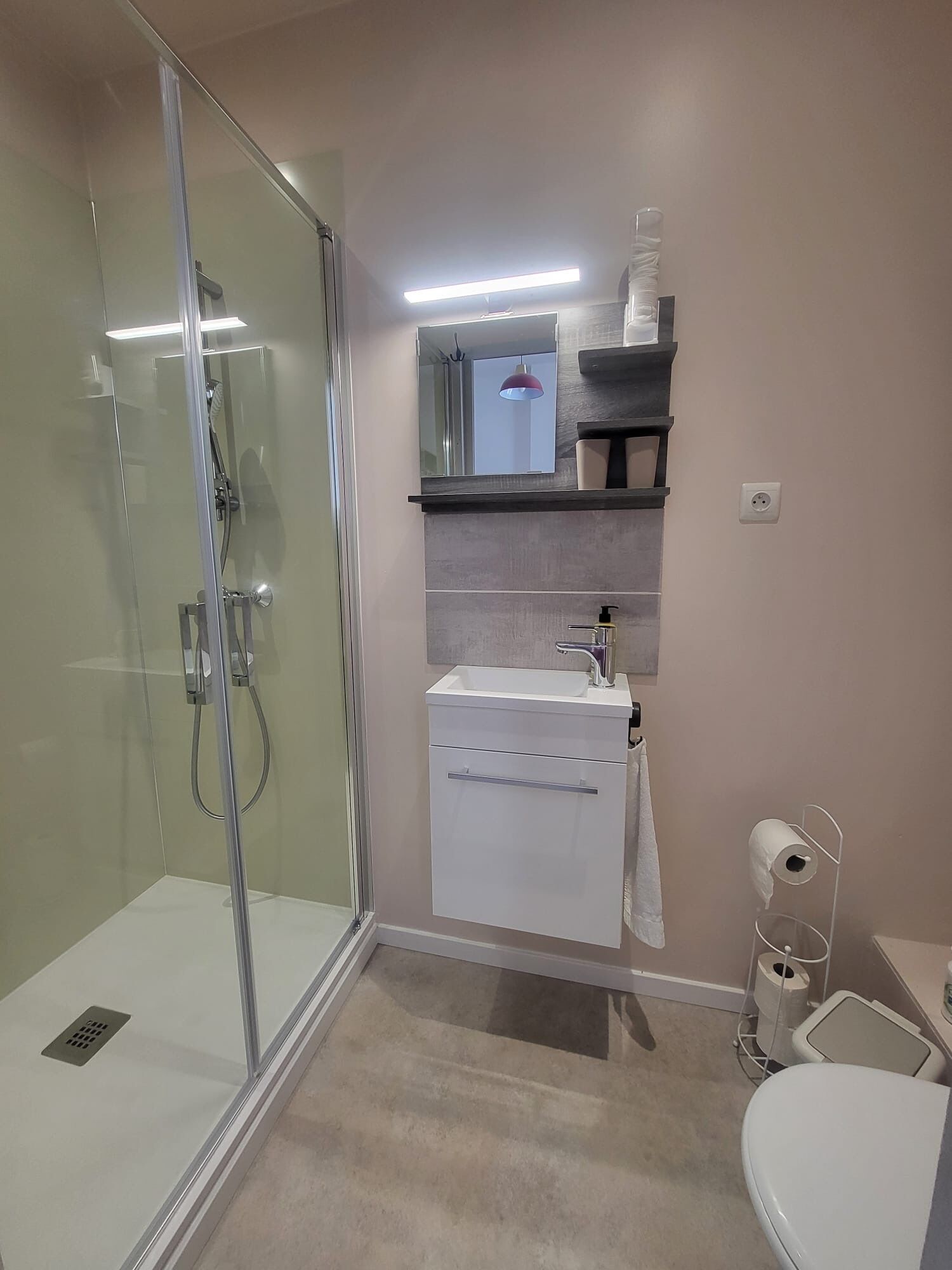 Une salle de bain moderne avec une douche à parois de verre, un lavabo blanc avec un miroir au-dessus et des toilettes dotées d’un porte-serviettes en papier.