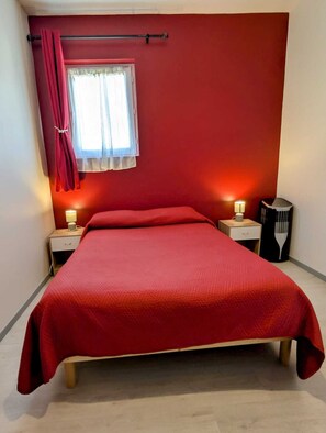 A bedroom with a red bedspread, a window with red curtains, two bedside tables with lamps, and a radiator. - Comme à la maison (Espéraza)