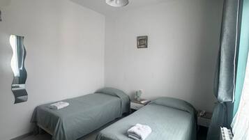 A hotel room with two single beds, a desk, a chair, a small table, a nightstand, a lamp, a picture on the wall, and a window with curtains.