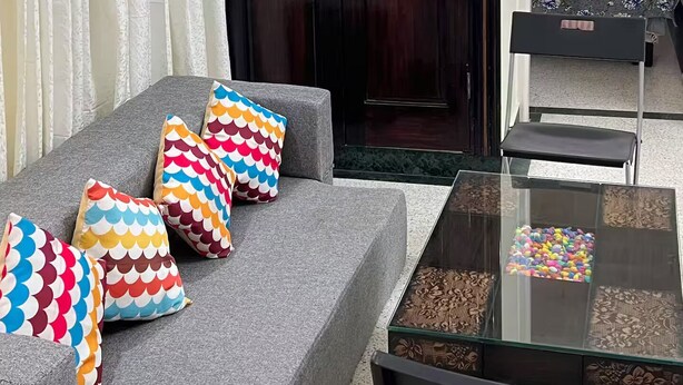 Een kamer met een grijze bank, kleurrijke kussens, een glazen salontafel, een houten garderobe en een bed met een bloemenprintdeken.
