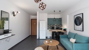 Ein modernes Wohnzimmer mit einem türkisfarbenen Sofa, einem runden Holztisch und einem gewebten Pouf. Im Hintergrund ist eine Küche mit türkisfarbener Spritzschutz und Pendelleuchten zu sehen.