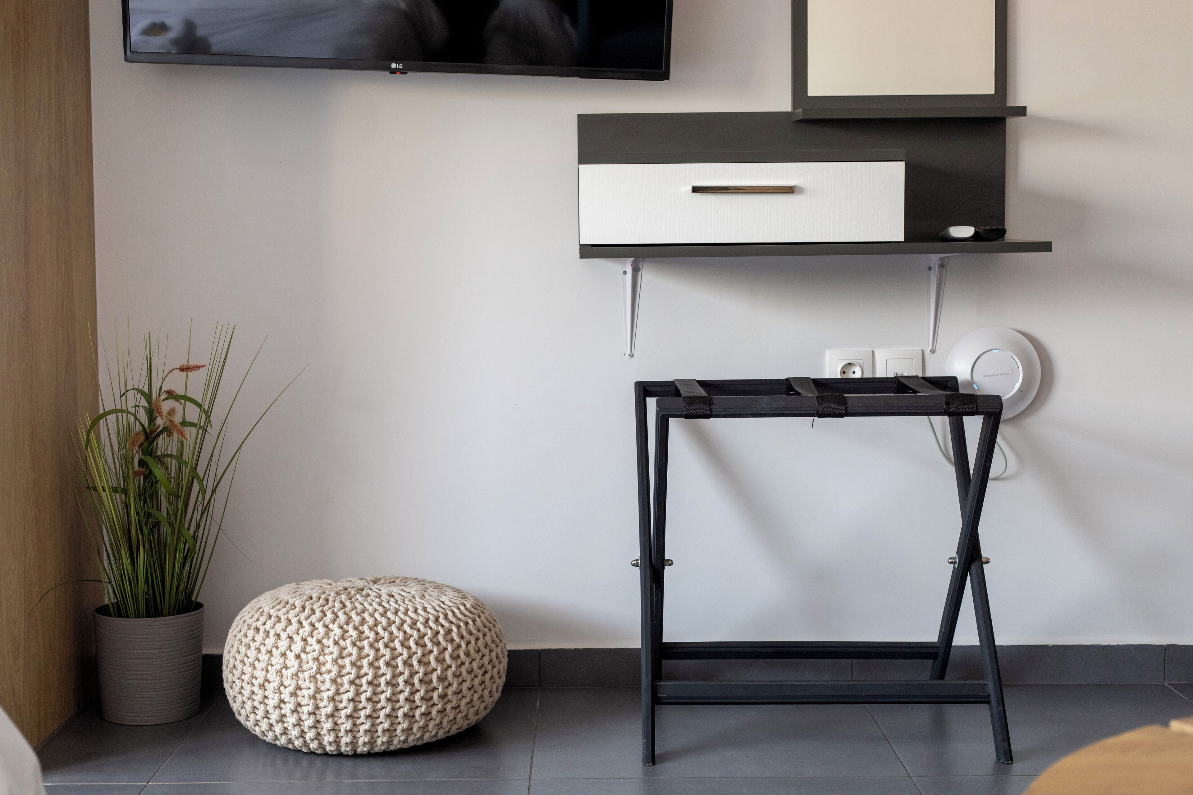 A modern room with a flat-screen TV mounted on the wall, a black desk with a drawer, a potted plant, and a round ottoman.