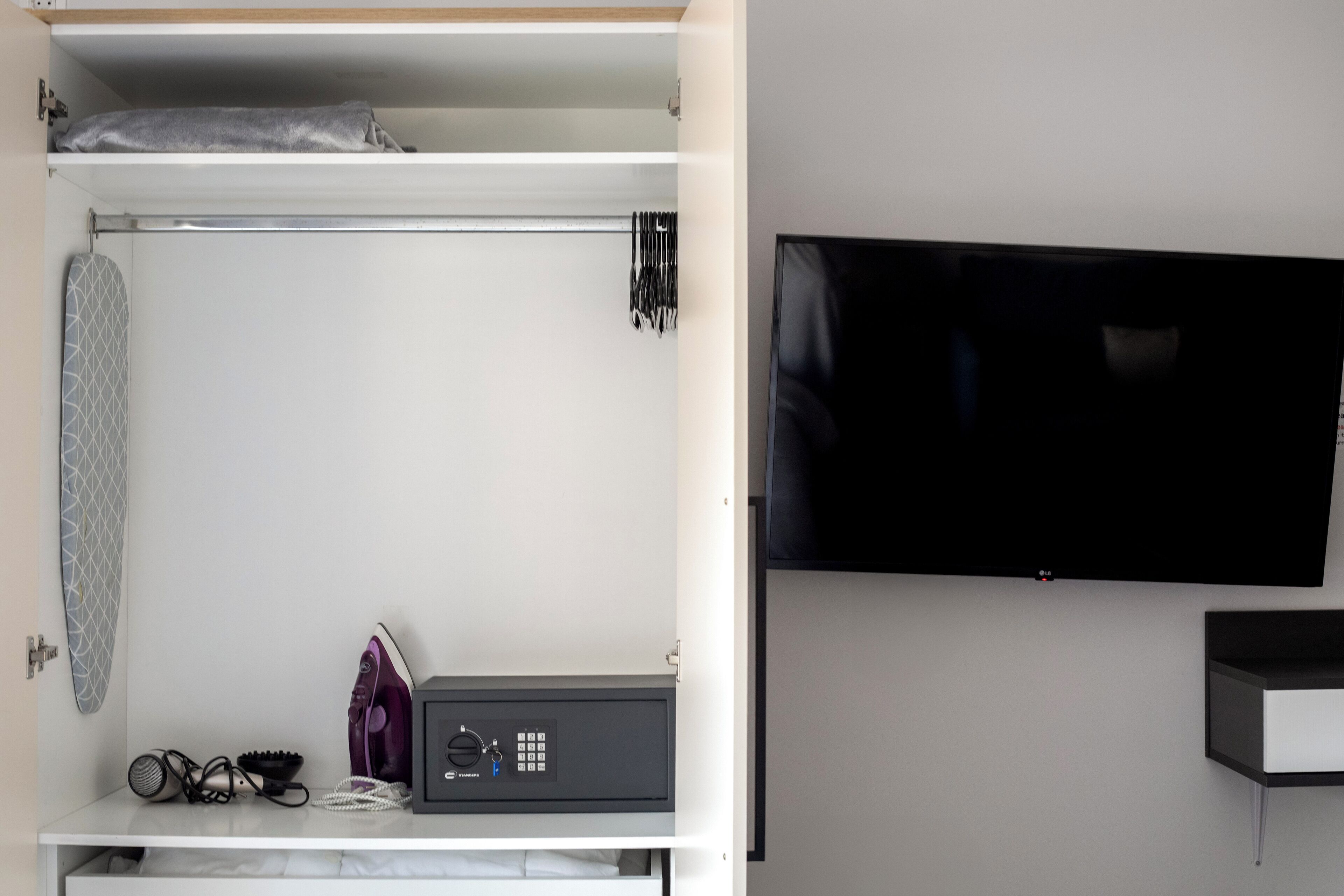 Un dormitorio con cama, televisor montado en la pared y una mesita con secador de pelo y otros objetos.