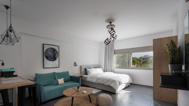 Ein modernes, helles Schlafzimmer mit einem Bett, einem petrolfarbenen Sofa, einem runden Couchtisch und einem großen Fenster mit Blick auf Grünflächen.