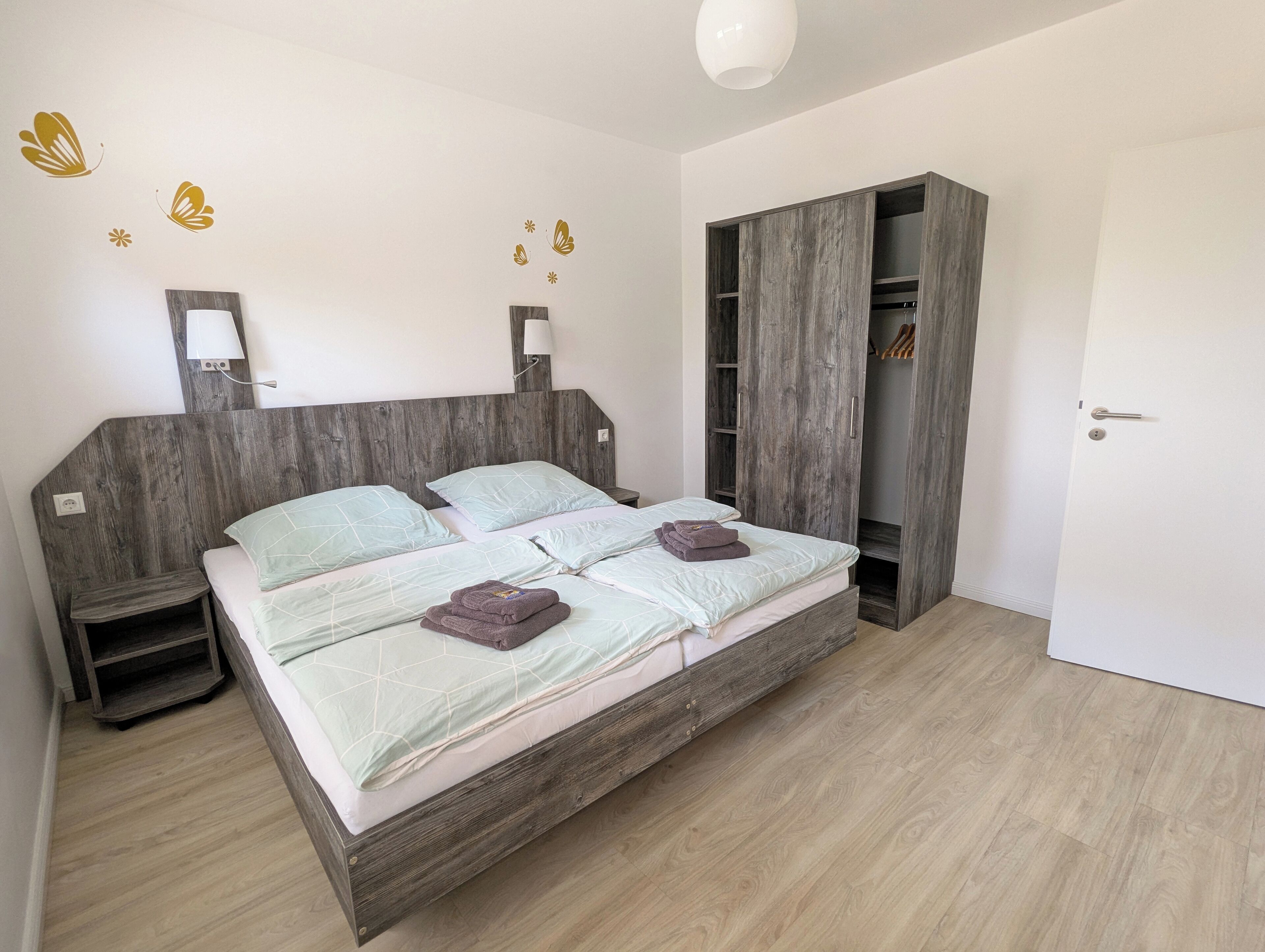 A bedroom with a wooden bed frame, two bedside tables, a wardrobe, and a wall-mounted lamp.
