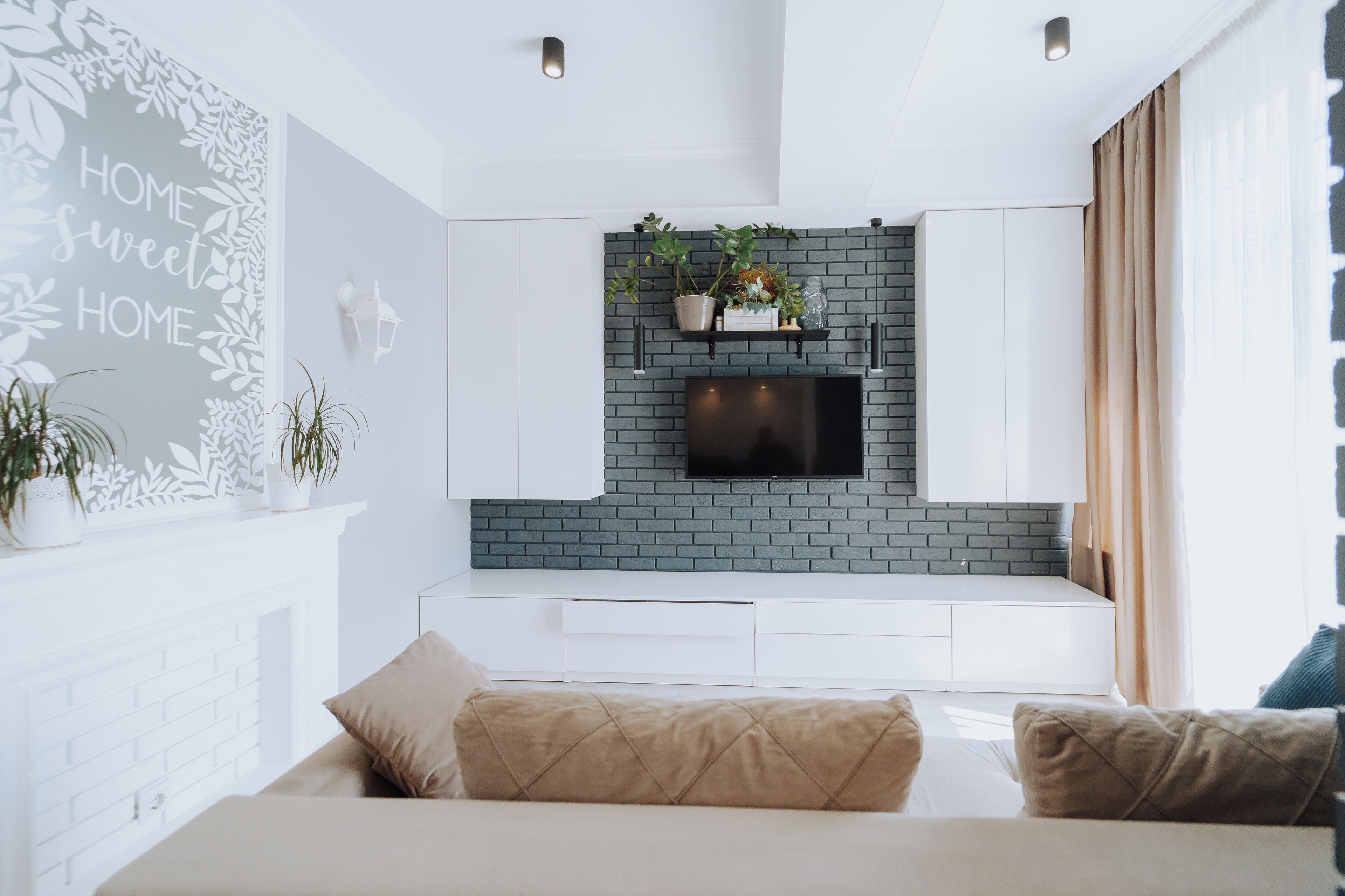 Una sala moderna con un sofá blanco, un televisor de pantalla plana montado en una pared de ladrillo, y una pared decorativa con la inscripción 'HOME sweet HOME'.