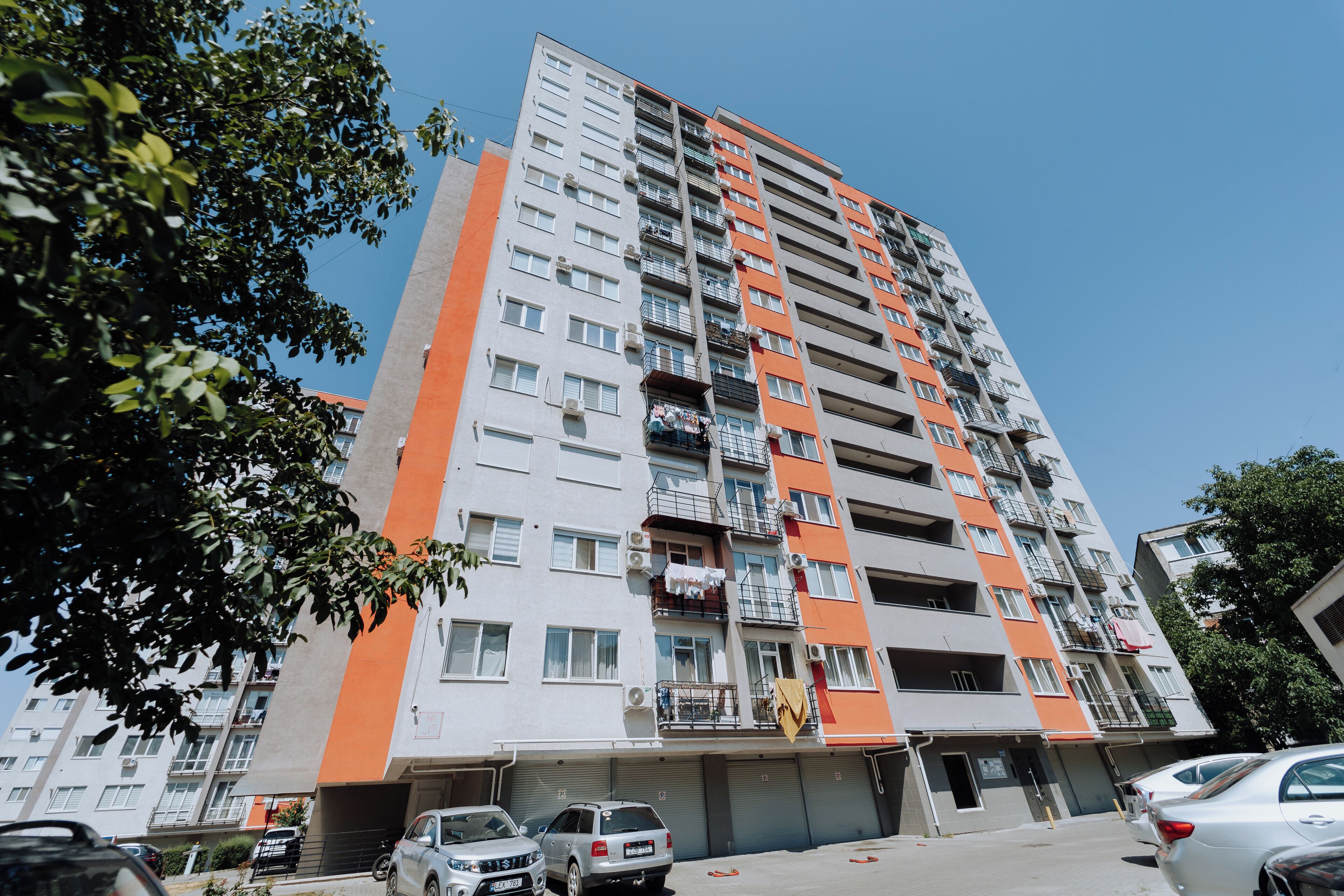 A multi-story residential building with balconies, a parking area, and a clear sky.