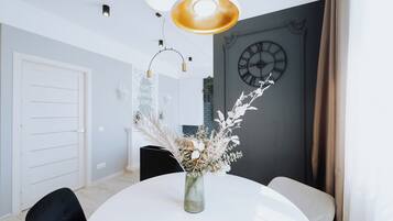 Uma sala de jantar com uma mesa redonda, cadeiras, um vaso com flores secas, luminárias pendentes, um relógio na parede e uma porta.
