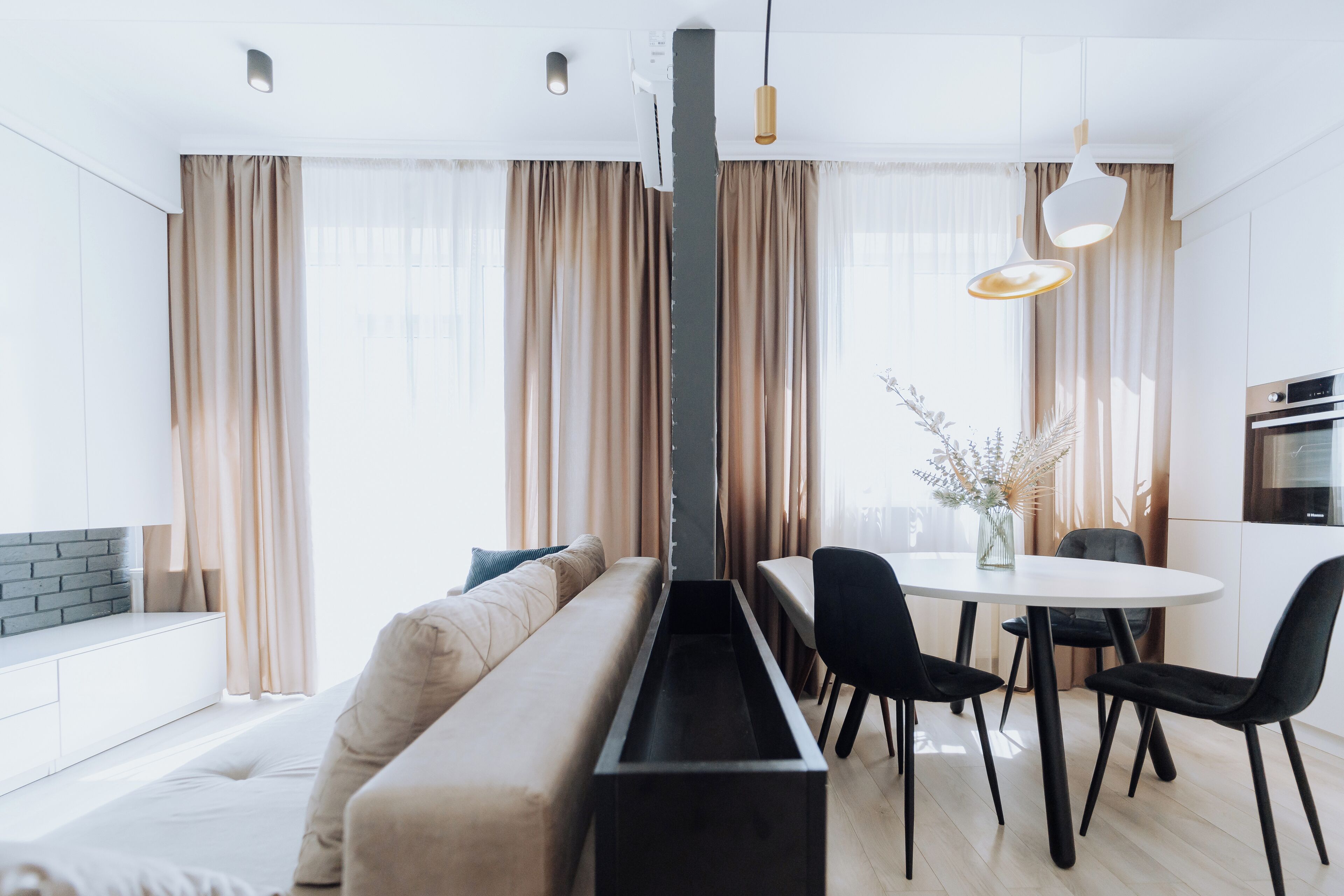 A modern living room with a sofa, dining table, and chairs. There is a kitchen area with an oven and a dining table with a vase of flowers.
