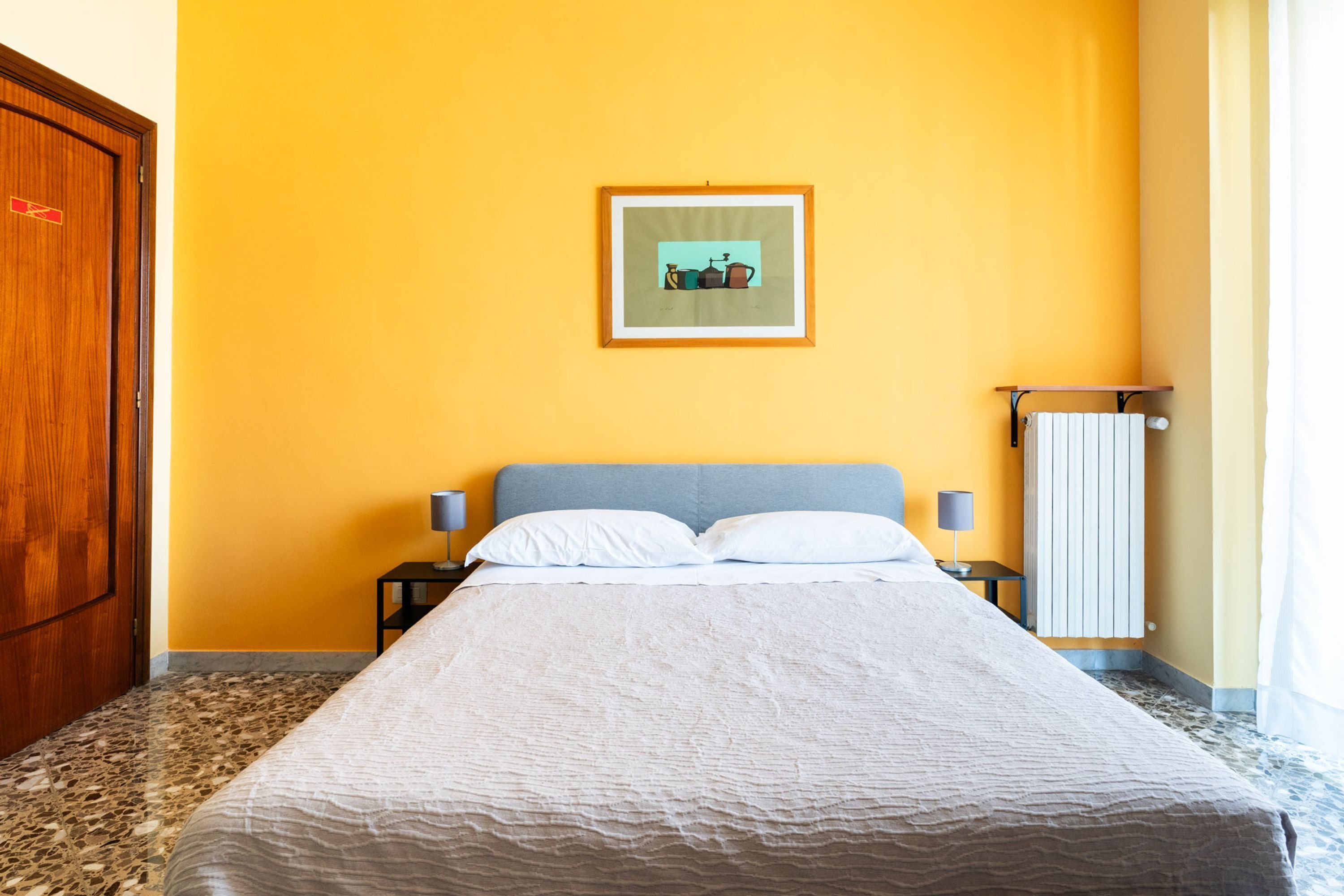 A hotel room with a bed, a framed picture on the wall, a nightstand, and a radiator.