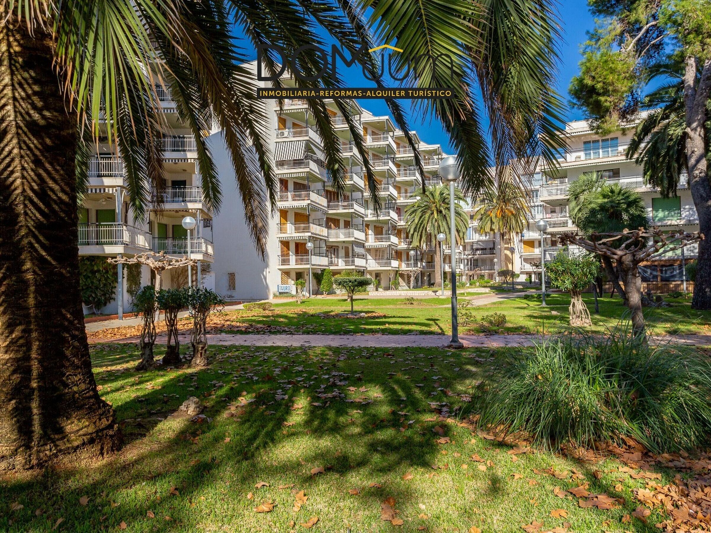 A multi-story residential building with balconies, surrounded by palm trees and a well-maintained lawn.