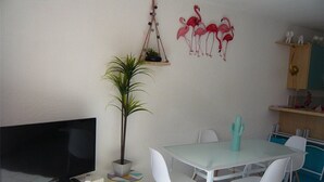 A modern living room with a flat-screen TV, a potted plant, a dining table with white chairs, and a wall-mounted shelf with decorative items. - Beautiful apartment with terrace for 4 people, near beach in Port Camargue (Port Camargue)