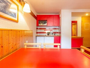 Une cuisine compacte avec des meubles rouges, un four à micro-ondes et un lave-vaisselle. On aperçoit un banc en bois et une table au premier plan.