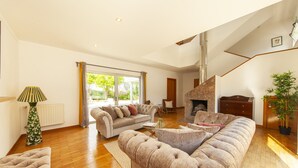 A spacious living room with a large sectional sofa, a smaller sofa, and a coffee table. There is a fireplace, a staircase, and a large window with curtains. - Villa Naturis (Portimão)