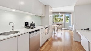 A modern kitchen with white cabinetry, a stainless steel sink, and a coffee machine. There is a dining area with a table and chairs, and a view of greenery outside. - The Sand Resort Kingscliff - Two Bed Dual Key (4215/16) (Tweed)