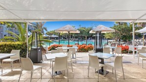 Un espace extérieur aménagé en terrasse, avec des tables et des chaises blanches, des parasols et une piscine en arrière-plan.