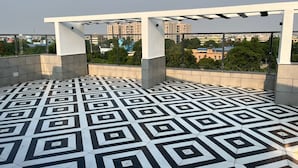 Eine Dachterrasse mit einem geometrisch gemusterten Boden, einer Pergola und Blick auf umliegende Gebäude und Grünflächen.