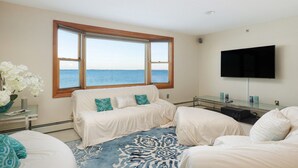 A room with a large window offering a sea view, a flat-screen TV mounted on the wall, and a blue patterned rug.
