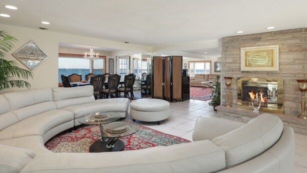 A spacious living room with a curved white sofa, a glass coffee table, and a fireplace. There is a dining area with a table and chairs in the background.