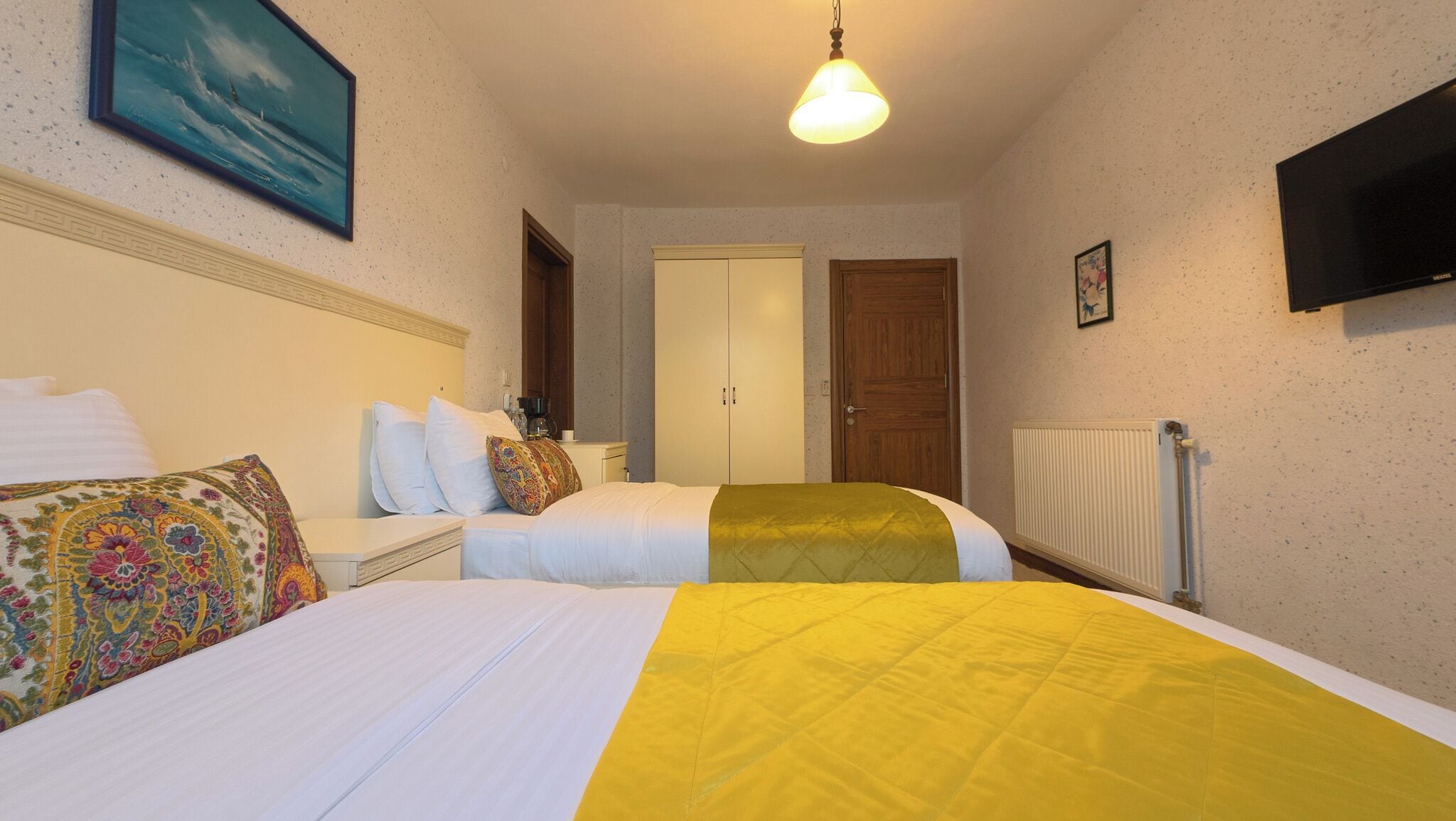 A hotel room with two beds, a television, a painting, and a radiator.