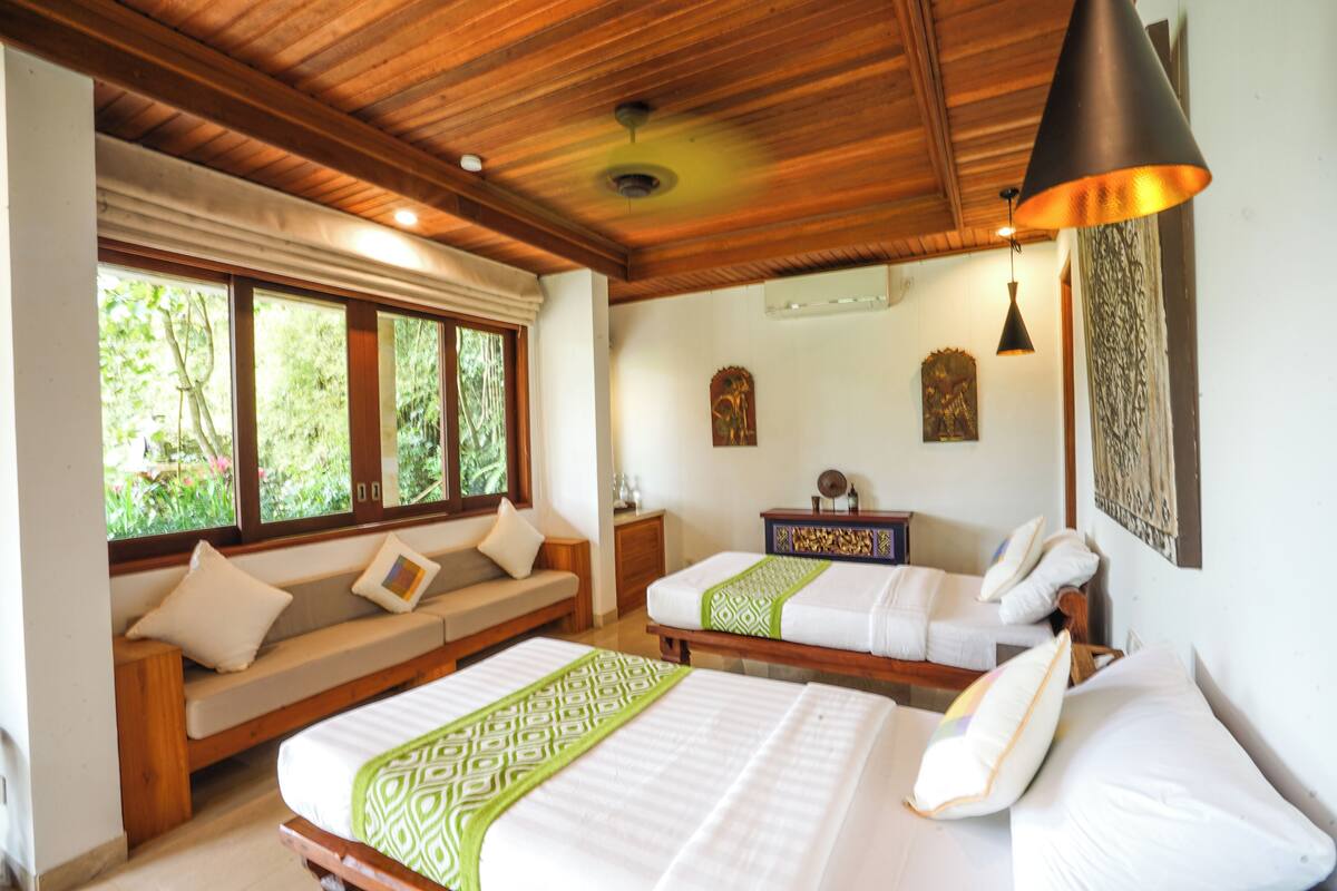 A room with two beds, a wooden ceiling, a window with a view of greenery, and a wall-mounted light fixture.