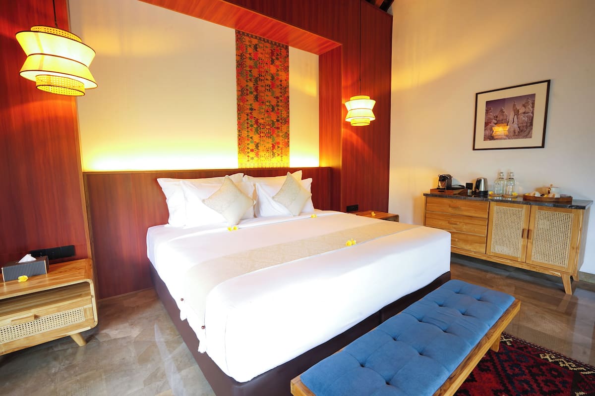 A hotel room with a large bed, bedside tables, a bench, a wooden dresser, and a wall with a decorative panel.
