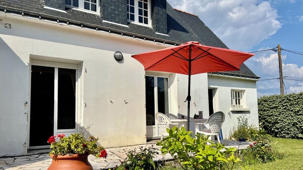 A house with a patio area featuring a red umbrella, white chairs, and potted plants. - La Roseraie-8P-Wifi-Jardin Clos (Quiberon)