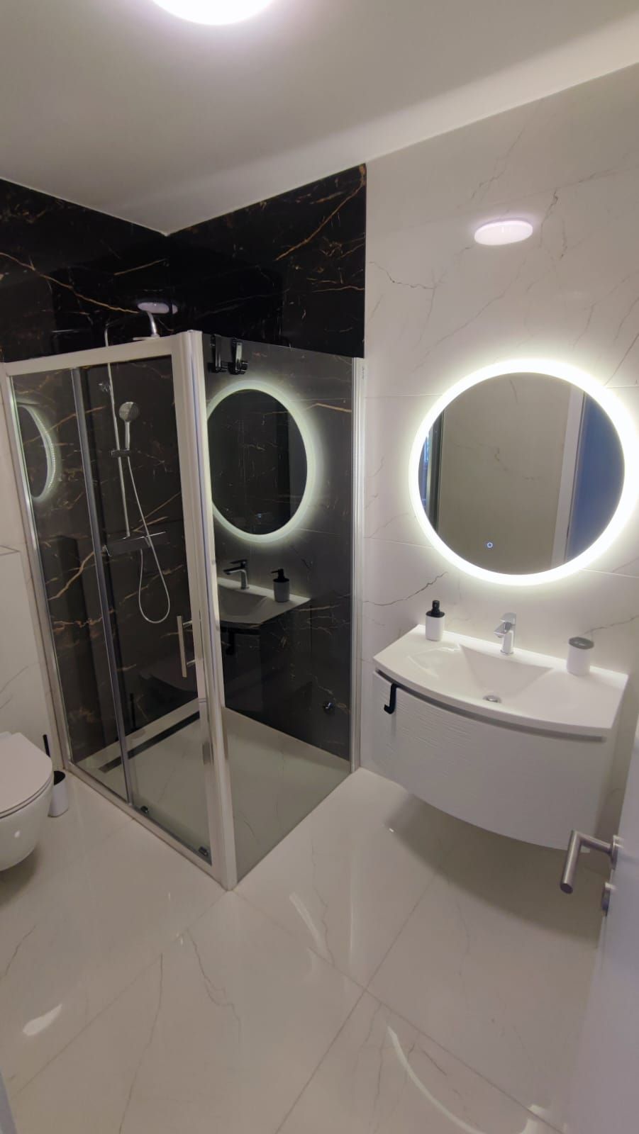 A modern bathroom with a glass shower enclosure, a round mirror, and a white sink.