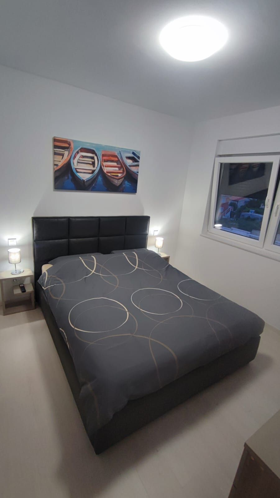 Un dormitorio con cama, dos mesitas de noche, una mesilla auxiliar, una ventana y un cuadro en la pared.