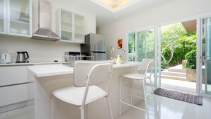A modern kitchen with white cabinetry, a stainless steel refrigerator, and a dining area with white chairs. - Tropical 3BR Pool Villa Morning Forest  (Rawai)