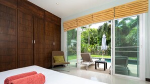 A bedroom with a large bed, wooden wardrobe, and a sliding glass door leading to a balcony with outdoor furniture. - Tropical 3BR Pool Villa Morning Forest  (Rawai)