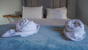 A bed with a grey headboard, white pillows, and a blue bedspread. There are two white towels on the bed. - A peaceful and safe area, just minutes from the beach, bars, and restaurants. (Cape Town)