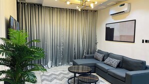 Un salon moderne avec un canapé d’angle, deux petites tables rondes, une grande plante en pot, un climatiseur fixé au mur et un luminaire suspendu au plafond.