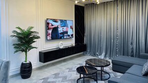 Un salon moderne avec un téléviseur à écran plat fixé au mur, une console noire, un canapé gris, deux petites tables rondes, une plante en pot et des rideaux gris.
