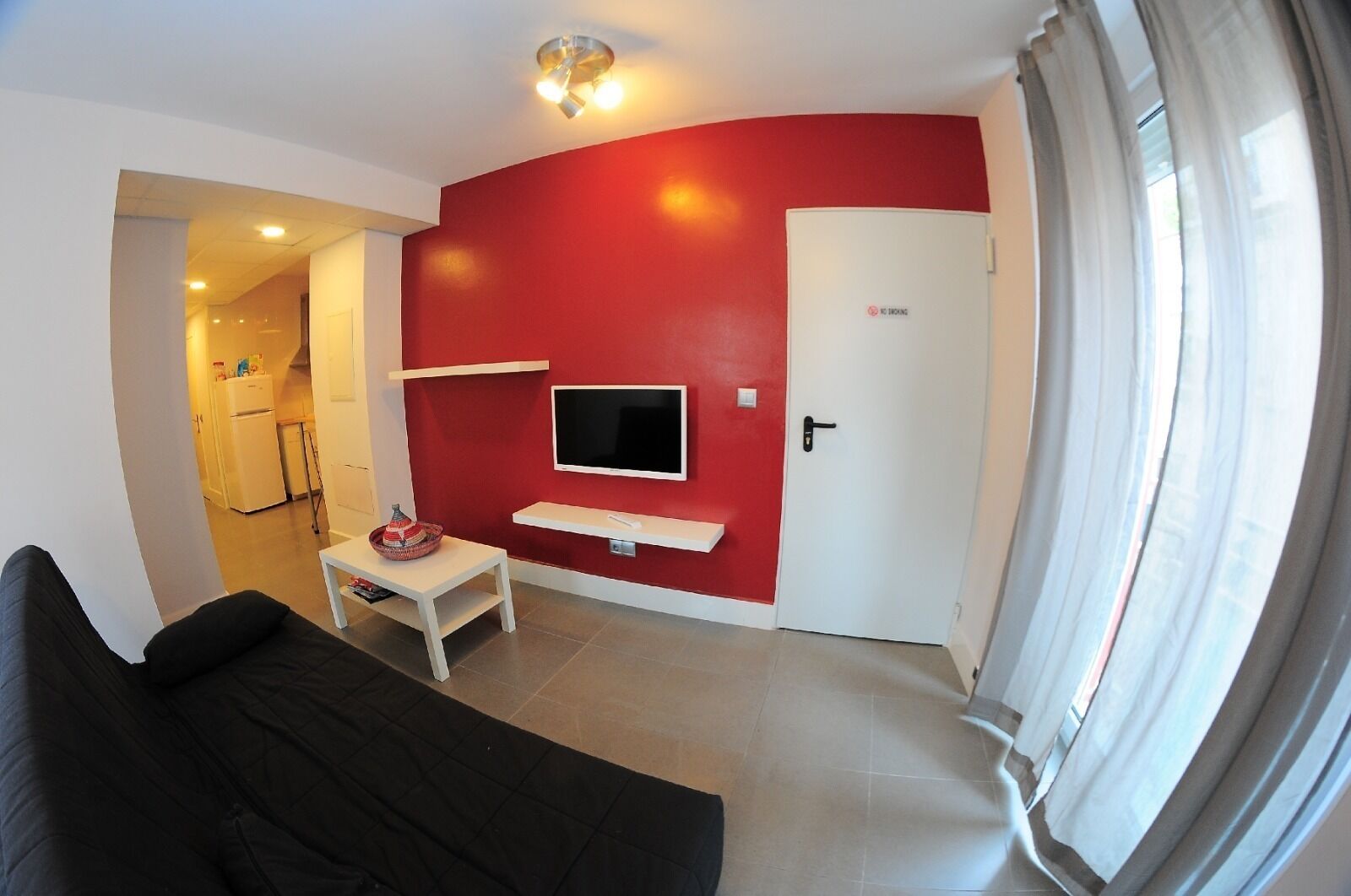 A living room with a red accent wall, a flat-screen TV mounted above a white console table, and a black sofa.
