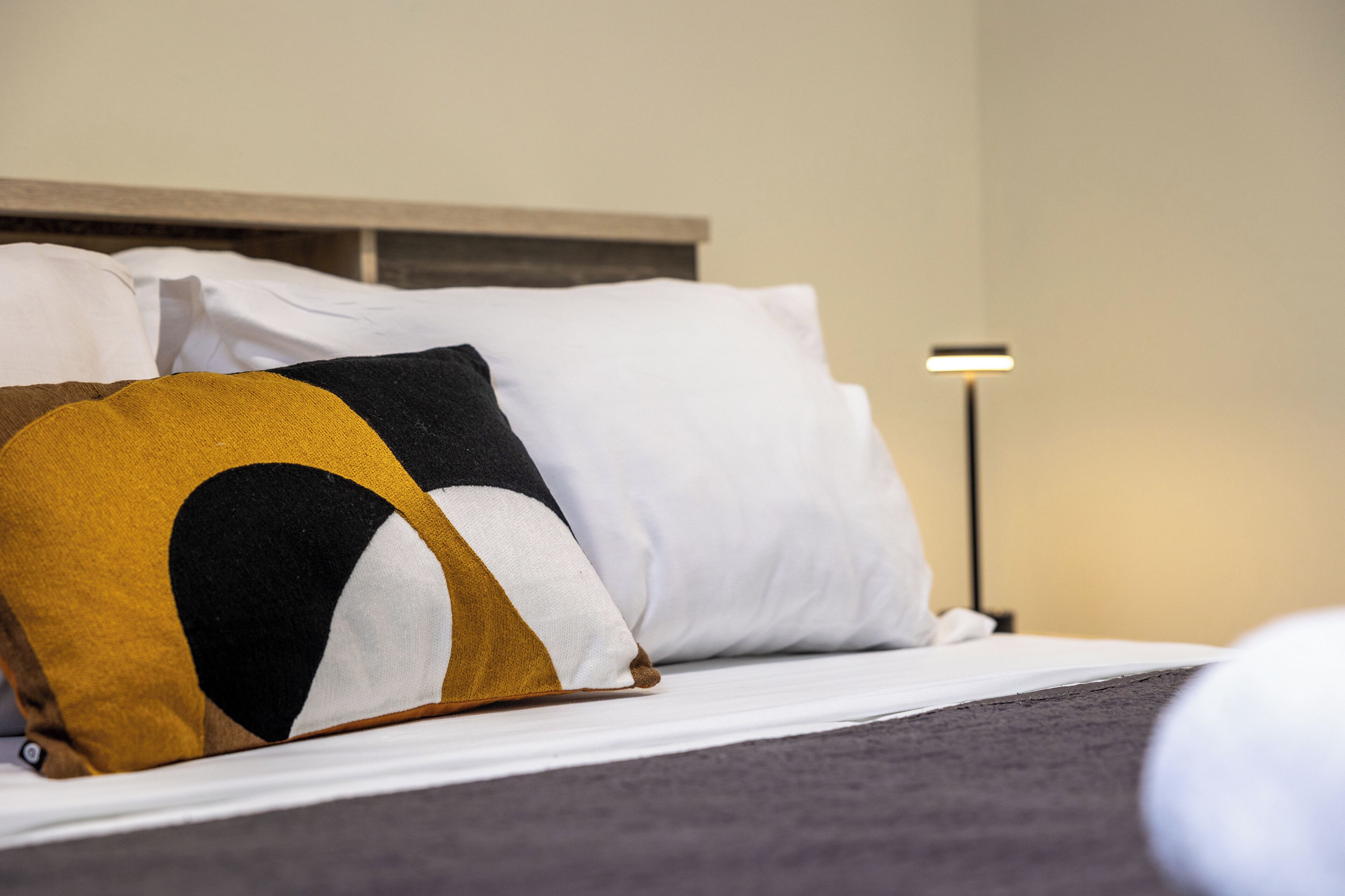 A bed with white and mustard pillows, a grey headboard, and a bedside lamp.