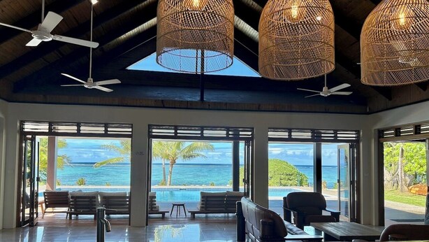 A spacious room with a large window overlooking a pool and ocean, featuring ceiling fans, wicker lamps, and a wooden table. - Mokusiga Villas Tolu - Private Beachfront Villa (Korolevu)