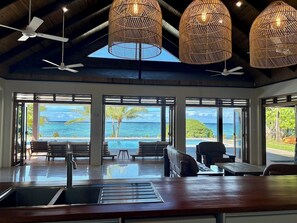 A spacious room with a large window overlooking a pool and ocean, featuring ceiling fans, wicker lamps, and a wooden table. - Mokusiga Villas Tolu - Private Beachfront Villa (Korolevu)