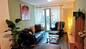 En opholdsstue med sofa, lænestole, et sofabord og et fjernsyn. Der er et stort vindue med gardiner, en plante og en lampe.