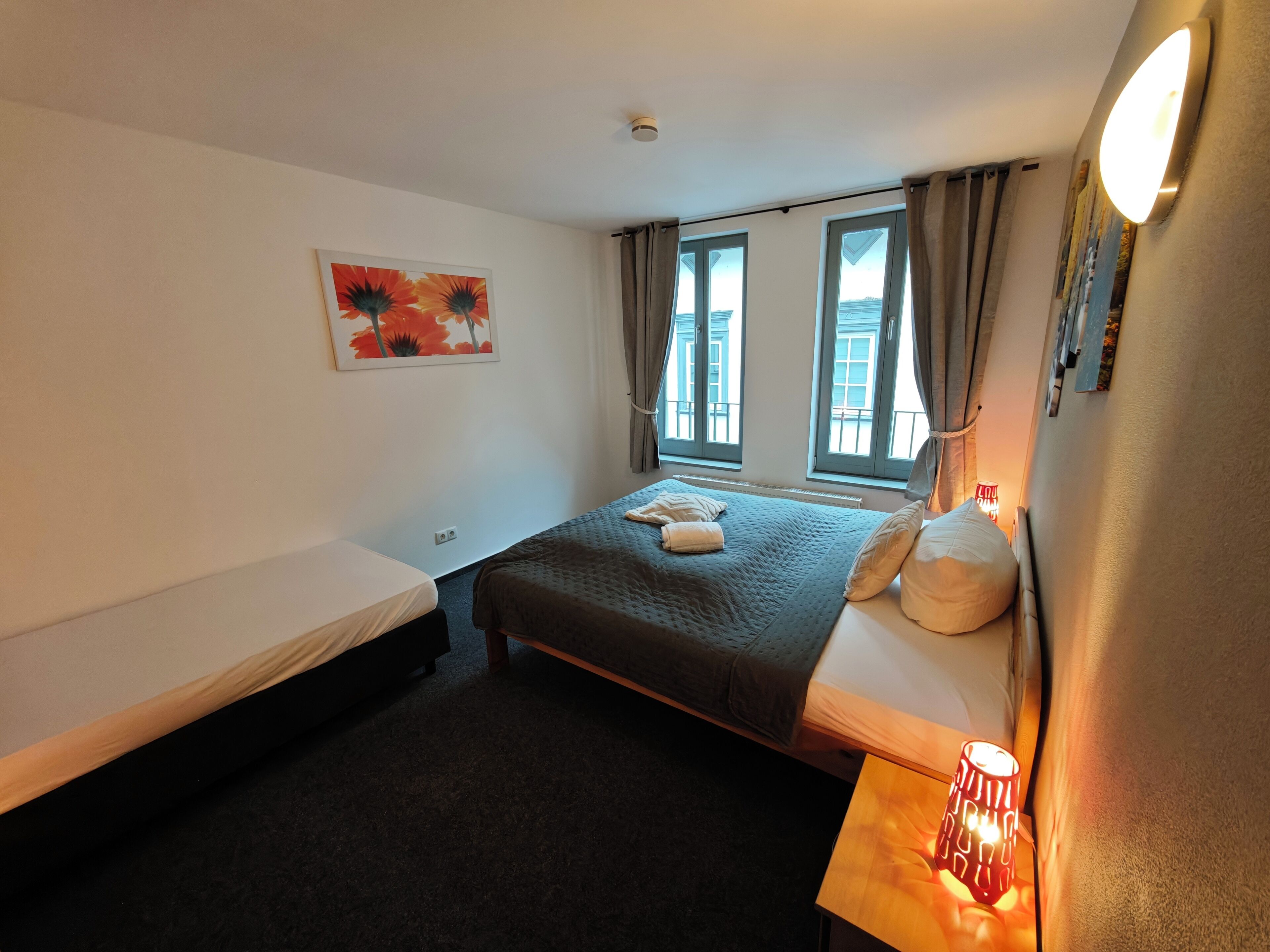 A hotel room with a single bed, a smaller bed, a nightstand with a lamp, a painting on the wall, and windows with curtains.