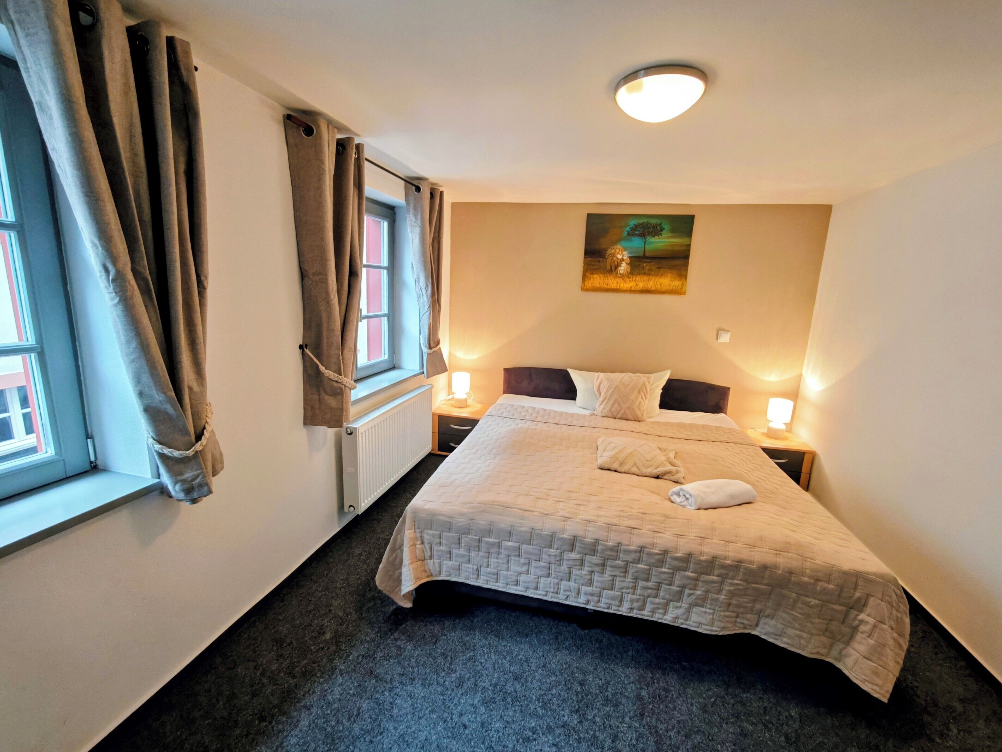 Ein ordentlich bezogenes Bett mit einer Decke, zwei Nachttischlampen, ein Fenster mit Vorhängen, eine Heizung und ein Bild an der Wand.