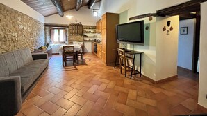 A spacious living area with a stone wall, wooden ceiling beams, a herringbone floor, a sofa, a dining table with chairs, a kitchen area, and a flat-screen TV mounted on the wall. - Locanda di Federico (Casteldelci)