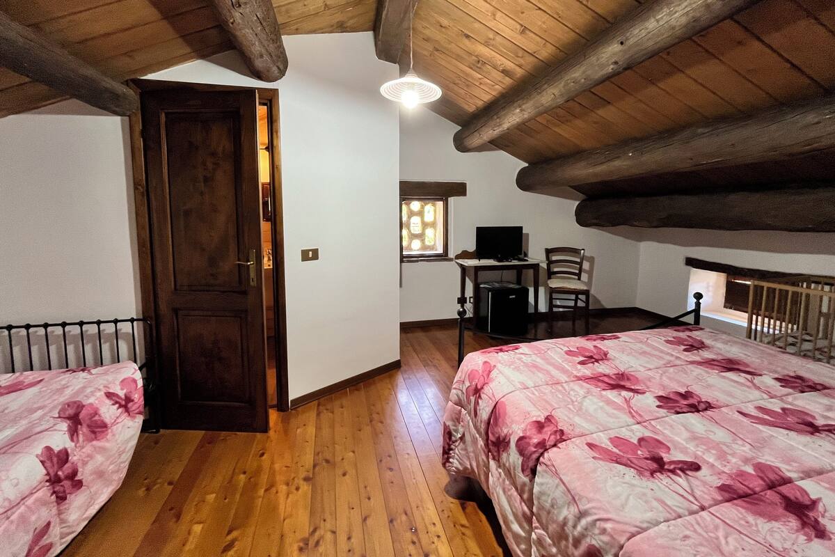 A bedroom with wooden beams, a bed with a floral bedspread, a desk with a computer, and a chair.