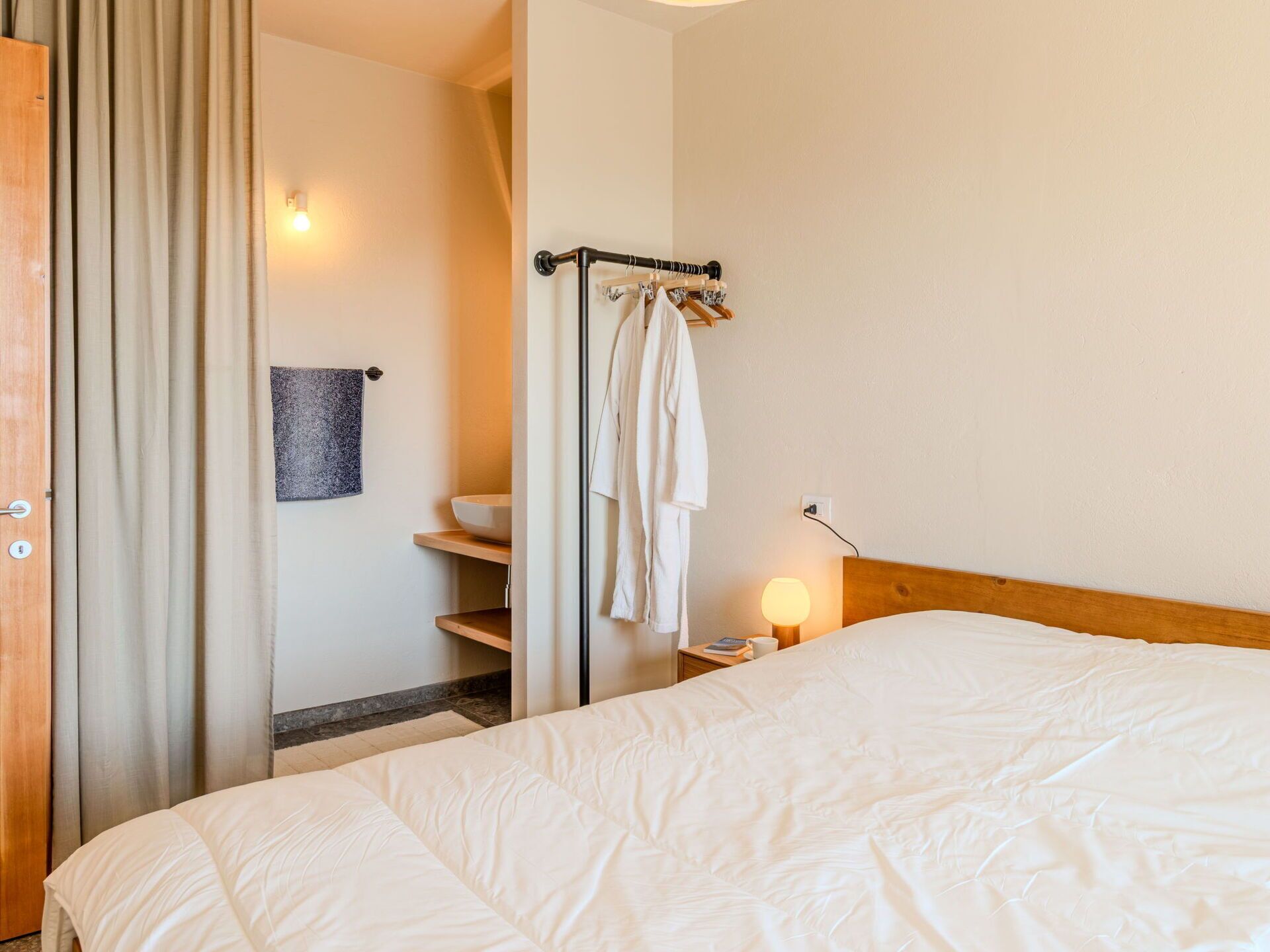 A hotel room with a bed, a nightstand with a lamp, a towel rack, and a bathroom visible through an open door.