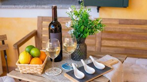 A wooden table with a bottle of white wine, two glasses, a basket of fruits, a vase with flowers, and white spoons on a placemat.