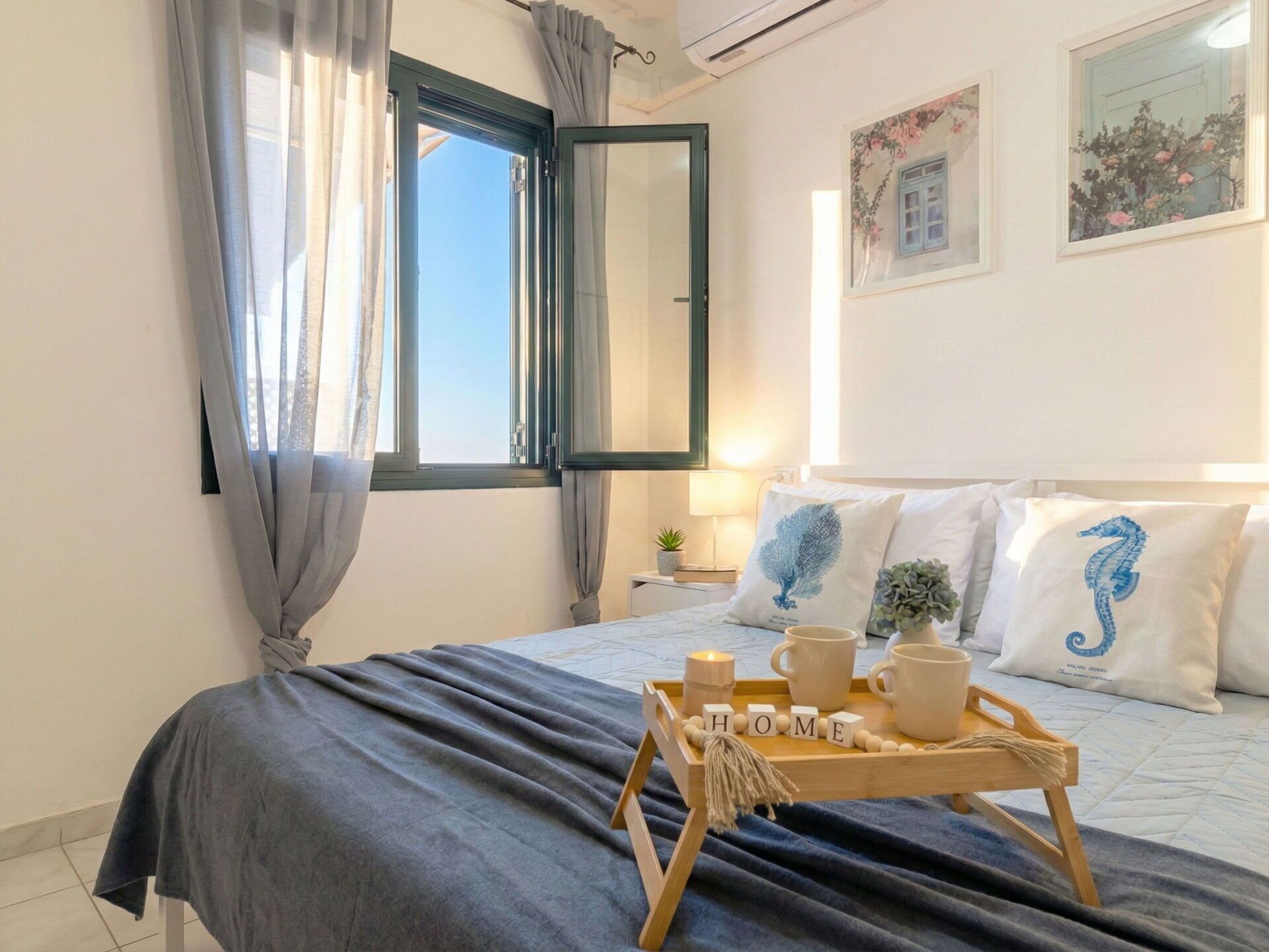 Een opgemaakt bed met een grijze sprei, een houten dienblad met kopjes en een bordje met de tekst 'HOME', en een raam met gordijnen.