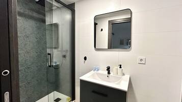 Un baño moderno con cabina de ducha de vidrio, un lavamanos blanco con espejo arriba y un mueble bajo en color oscuro.